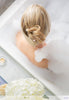 Blonde model in a candle lit bubble bath with hydrangea flowers on the ledge of the tub. The back of her head and shoulders are above the water with bubbles covering her right shoulder. Her hair is held in an all-up style by a bow and heart rose colored Flexi Clip. 