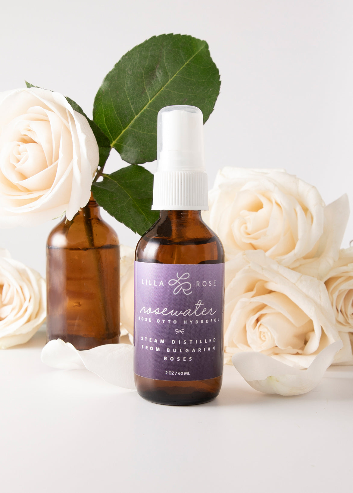 Rosewater in an amber glass bottle with purple lilla Rose label in front of white roses 