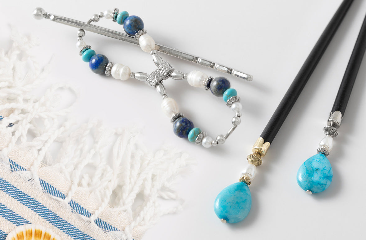 A pair of black hair sticks with turquoise blue stone beads and silver-toned metal accents, displayed on a white surface next to a white lace textile.